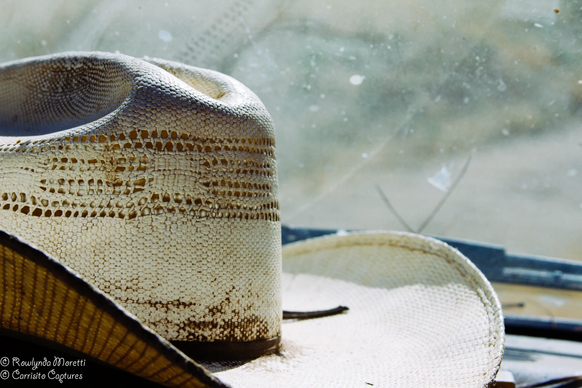 A pale straw cowboy hat with a vented crown rests on the dashboard of a vehicle, lit hard from the side, seen against a dusty windshield.