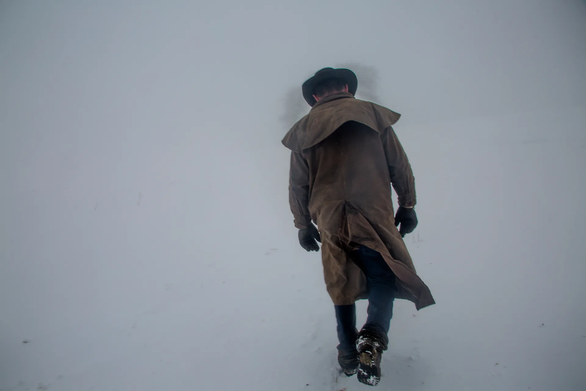 A rancher in a long oilskin duster, felt hat and gloves walks away from the camera into a whiteout, boots breaking through fresh snow.