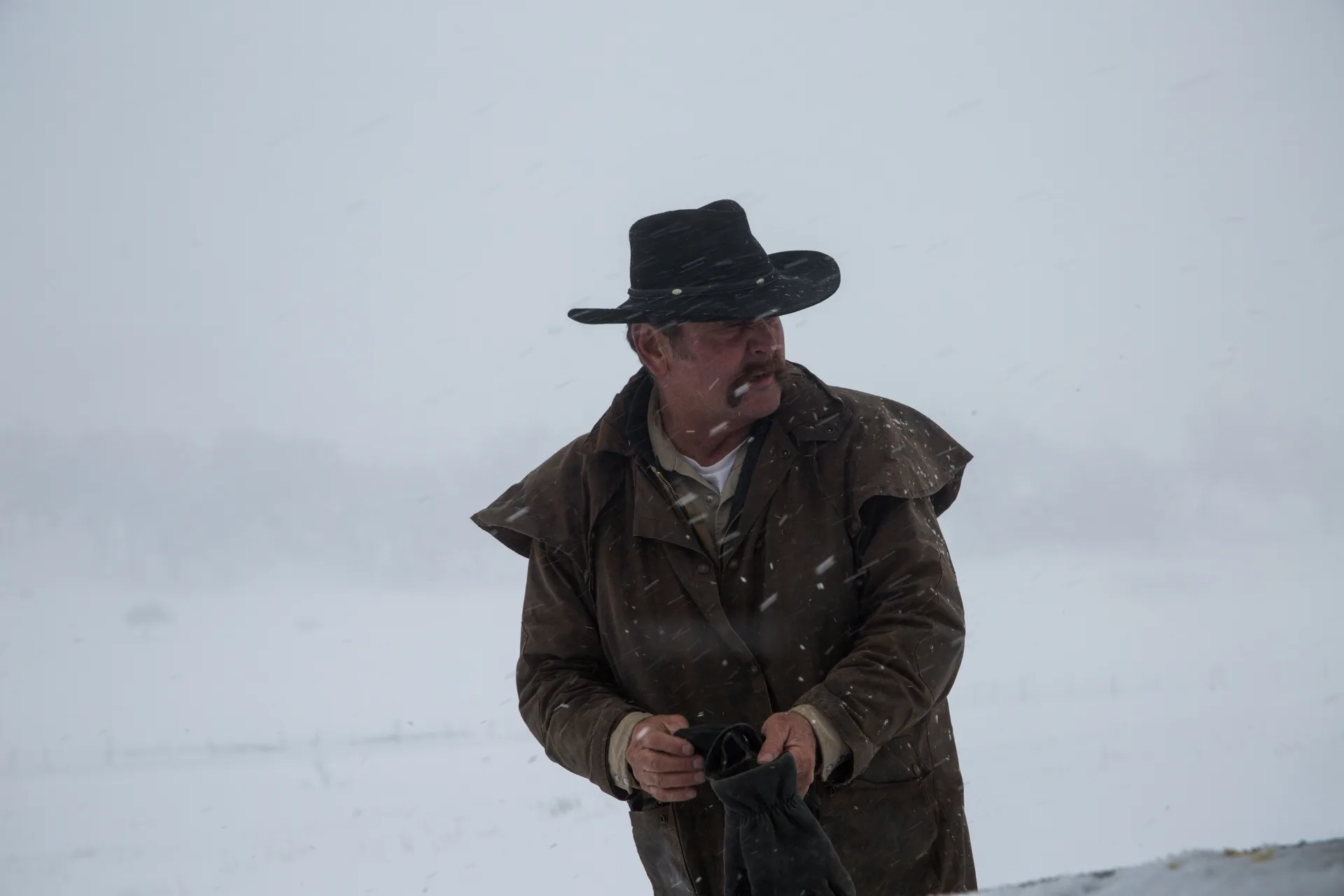 A rancher in a black felt hat and brown oilskin coat stands in falling snow pulling on his work gloves, looking off to his left, an open white field and a wire fence behind him.